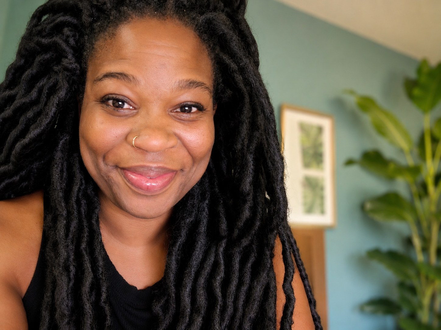 Charisse Fontes, founder and host of Sticks & Stones Crystal Tours, smiling warmly in a green-walled room with plants and framed botanical prints
